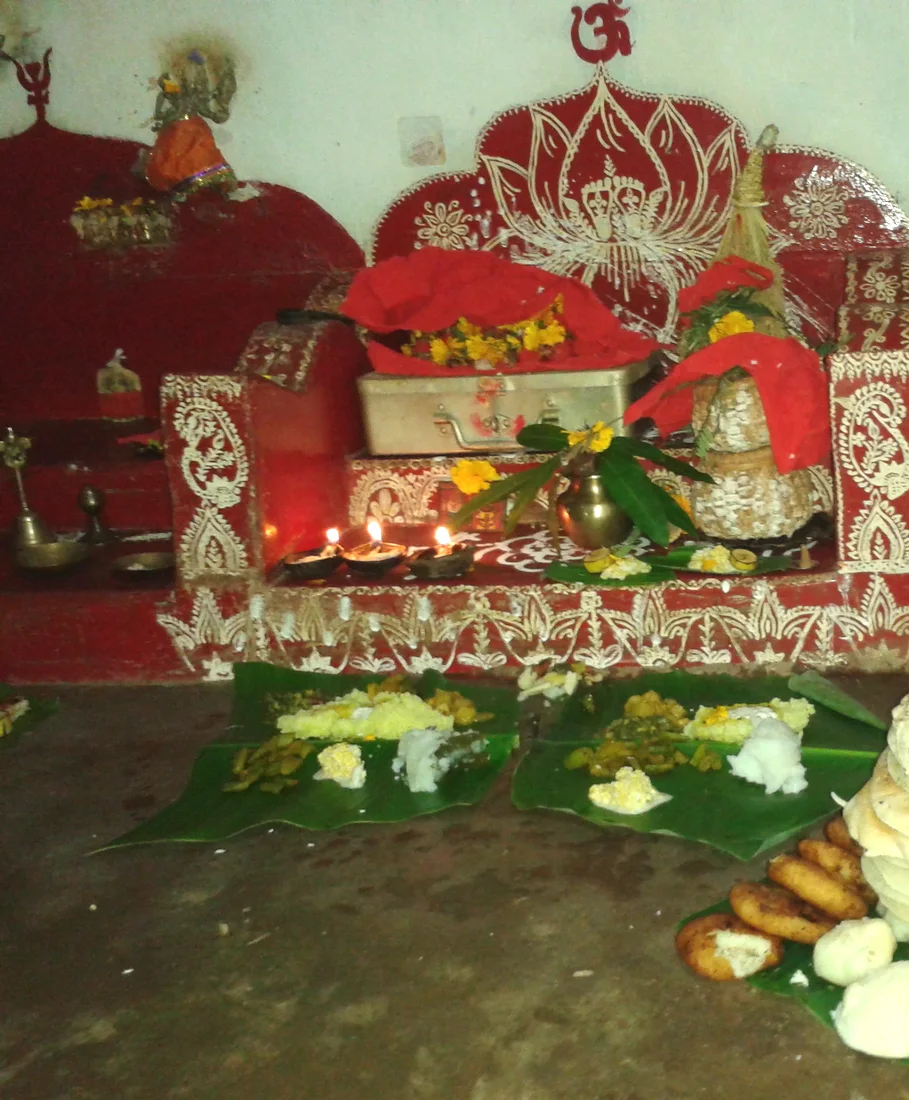 A Manabasa Gurubara observance in an Odia home — the household shrine painted with jhoti in rice paste, the row of lamps lit, and the offerings and pithas of the new crop set out on banana leaves