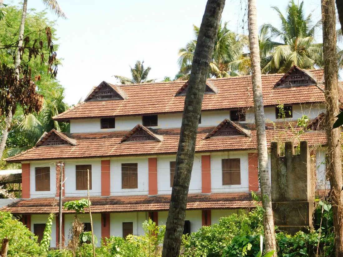 A traditional Kerala nalukettu taravad house with tiled roofs and carved wooden gables among coconut palms, the ancestral home of a Nair family