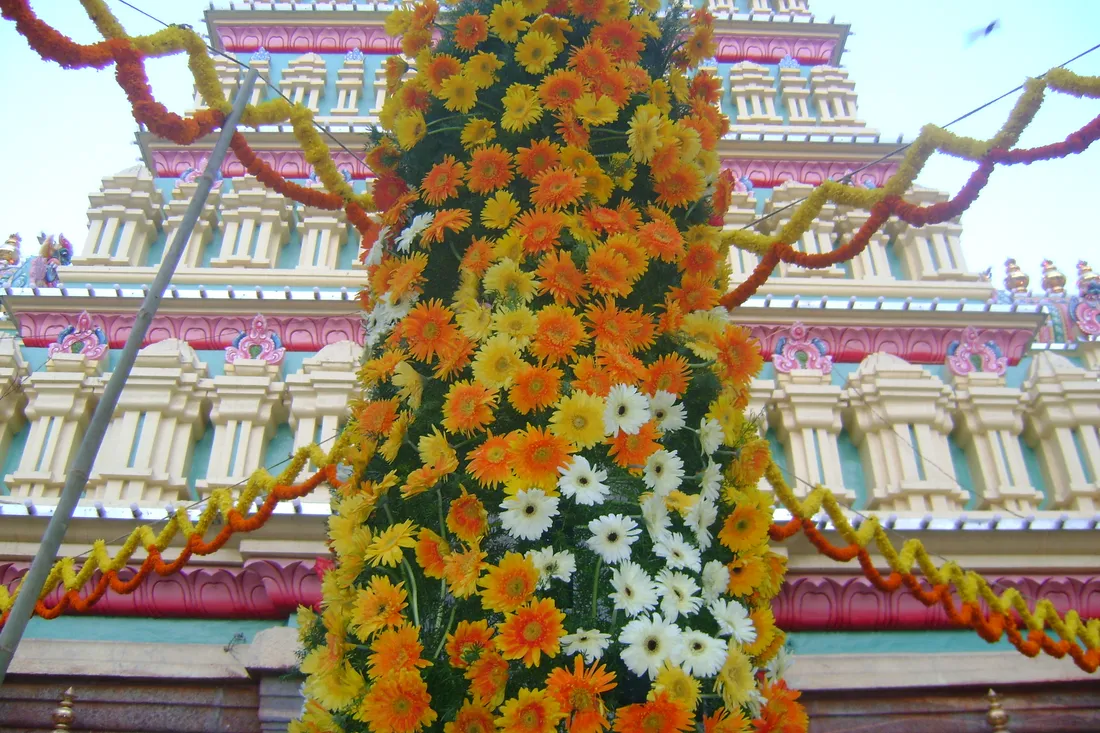A tall column of orange, yellow and white gerberas raised against the painted gopuram of the Dharmaraya Swamy temple, with marigold garlands strung across the front, on the day before the Bengaluru Karaga